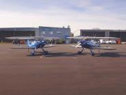 Two bi-planes parked outside with hangar buildings in background