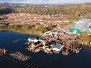 Dock with boathouse on river, land with log yard, industrial buildings, trees in background