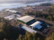 Aerial of river, log yard, industrial buildings with parking and packaged product nearby, trees and sky