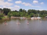 Six people on stand up paddle boards in river, marina with boats in background, trees, sky